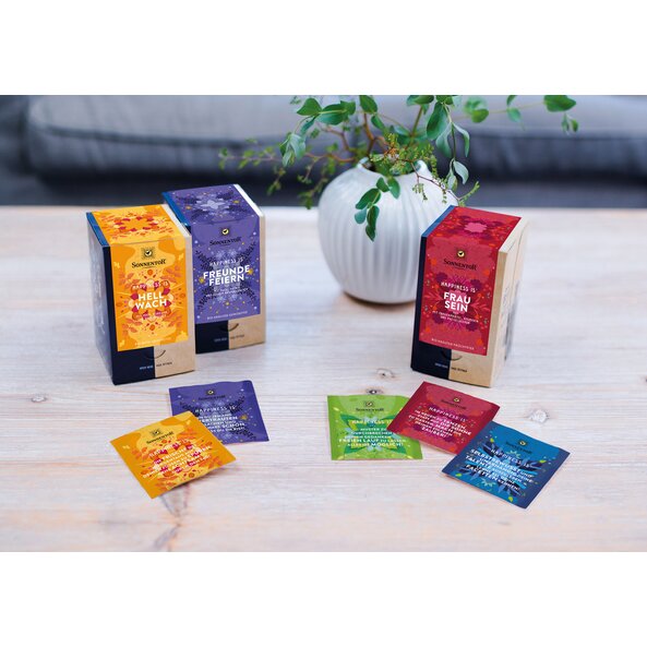 Auf dem Bild sind drei Happiness Is Teepackungen in den Farben lila, rot und gelb zu sehen. Diese stehen auf einem Tisch. Dahinter ist eine Pflanze zu sehen.